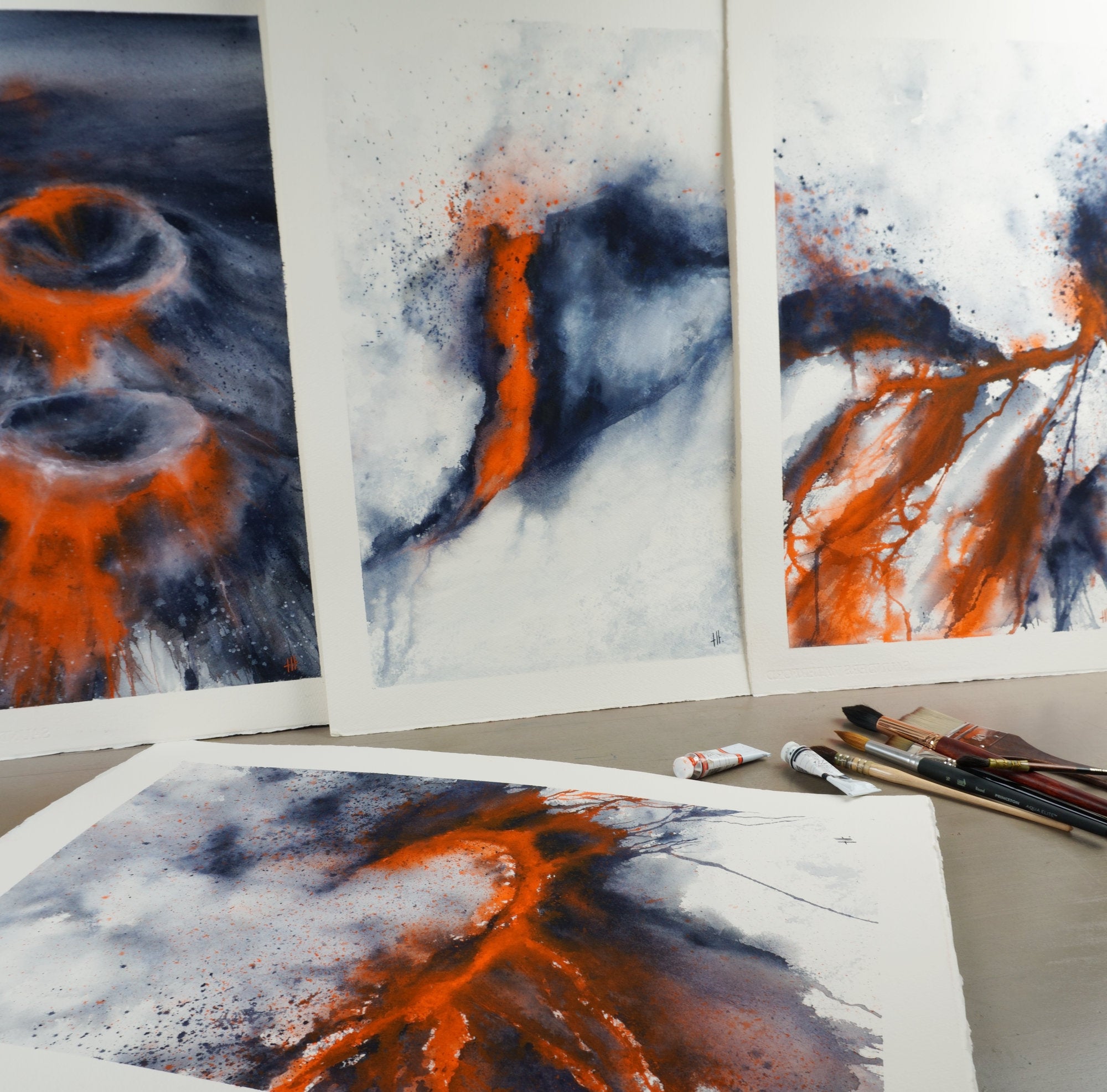 Upright and flat studio display of Rings of Fire, Red Zone, River of Death and At 1000 Degrees, part of The Magma Collection by Haidi Hildemore, alongside brushes and M. Graham watercolour paints.